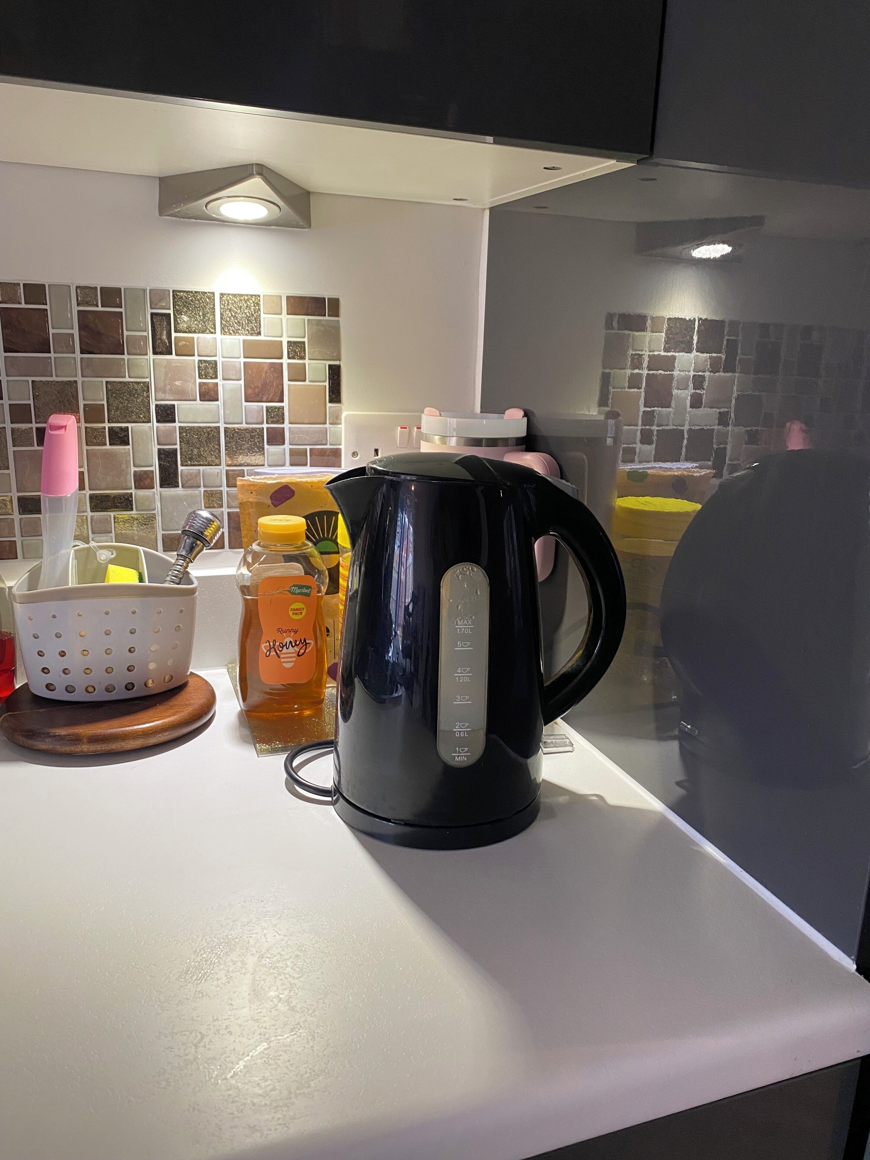 A kitchen counter with a black electric kettle, a colander, a jar of honey, and a bottle of honey.