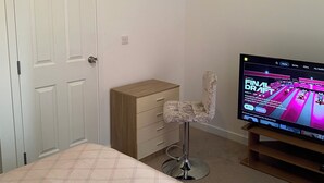 Une chambre à coucher comprenant un lit, une commode en bois, une chaise, un téléviseur et une porte avec un porte-manteau.