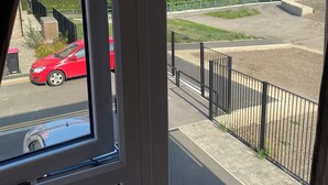 Une vue depuis une fenêtre donnant sur un parking où se trouve une voiture rouge, un bâtiment à la façade en briques et un espace vert clôturé.