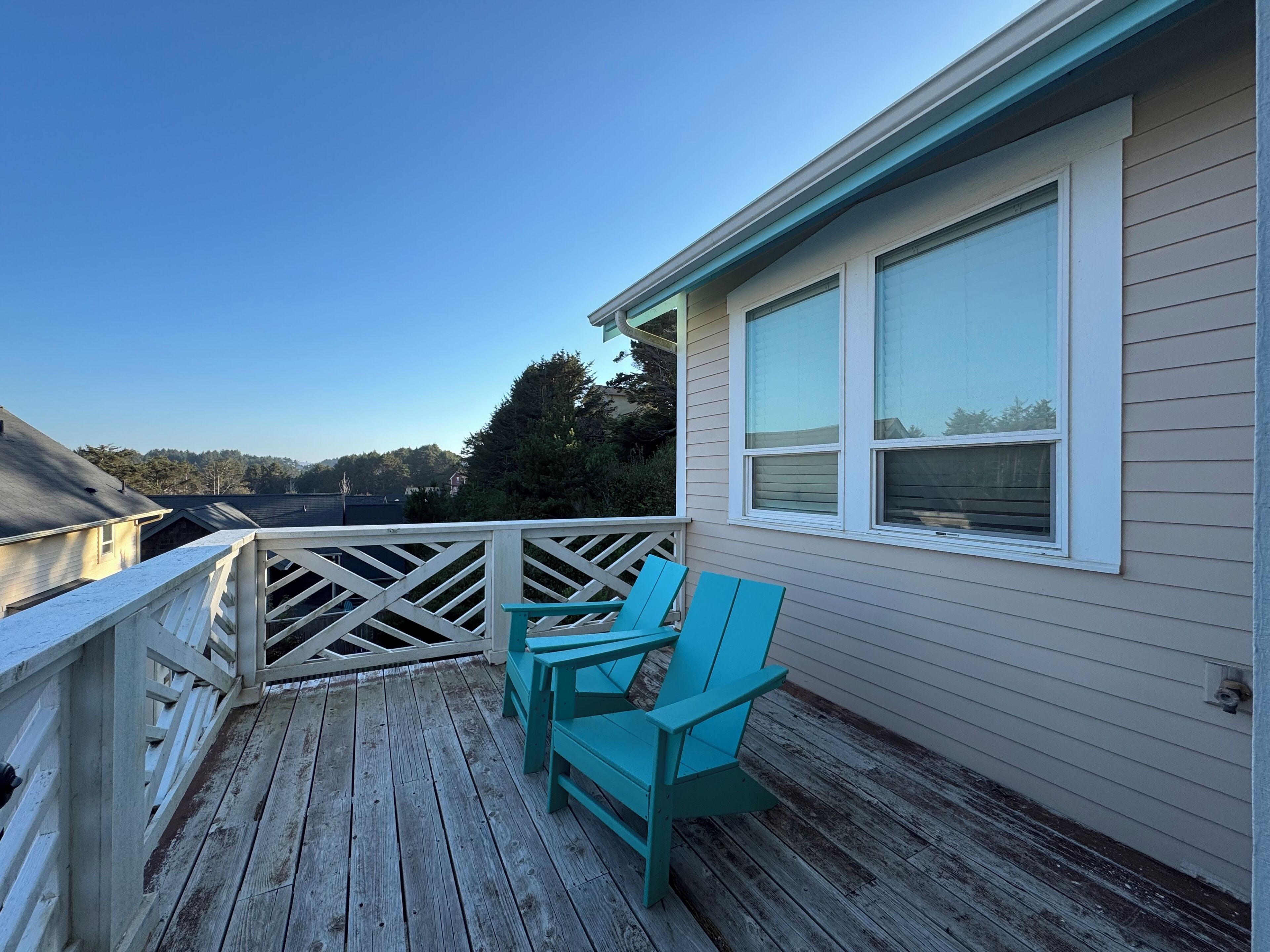 Una terraza de madera con sillas turquesa y barandilla blanca, con vista a una zona residencial con casas y árboles.