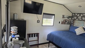 A bedroom with a bed, a TV mounted on the wall, a small table with a basket, a chair, and a window. - Cozy Cottage With a View in La Grange (La Grange)