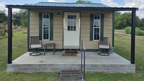 A small, single-story building with a green door and windows, a covered porch with two chairs and a table, and a metal chair in front. - Cozy Cottage With a View in La Grange (La Grange)