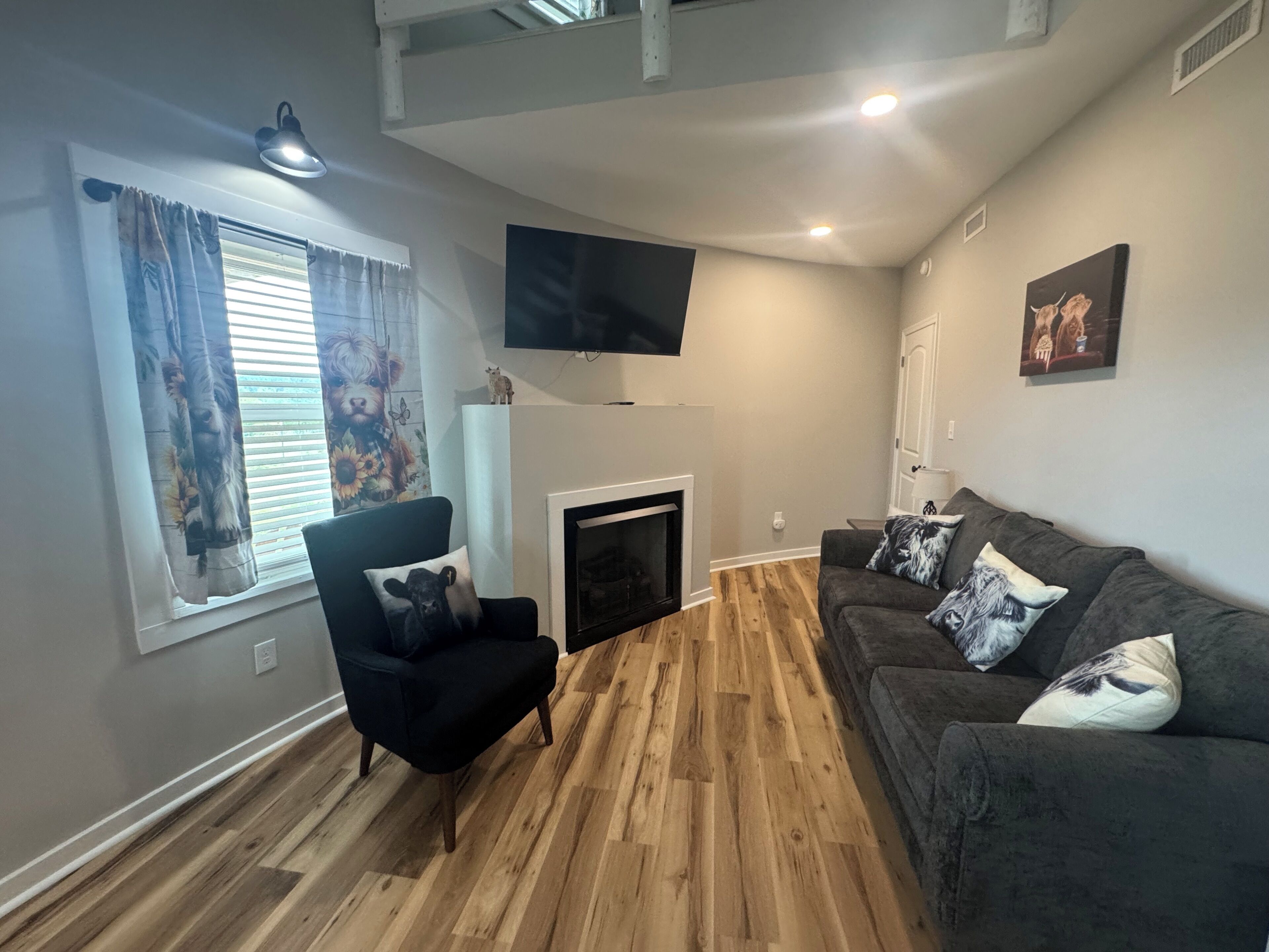 Una sala de estar con chimenea, un sillón negro, un sofá gris, piso de madera, televisor montado y una ventana con cortinas.