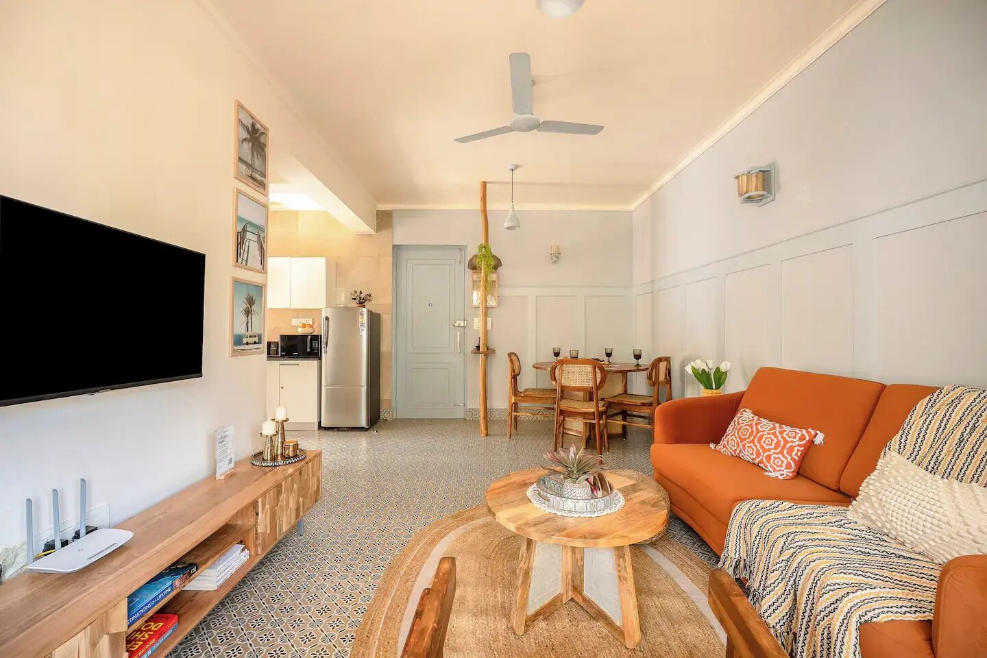 A modern living room with a flat-screen TV, a wooden coffee table, an orange sofa, and a dining area with a chandelier.