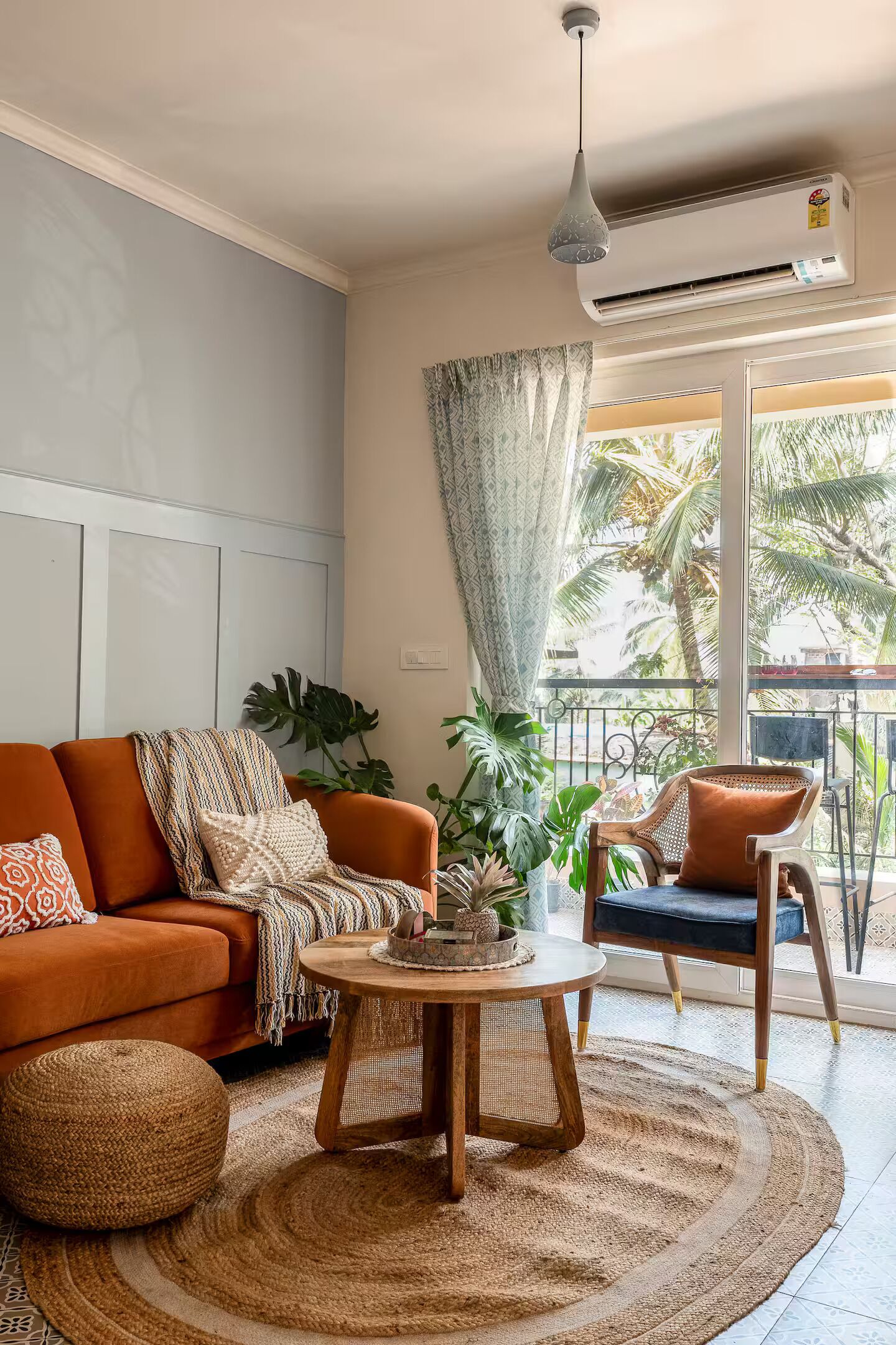 Een woonkamer met een bank, een rond bijzettafeltje en een stoel. Er is een raam met gordijnen, een plant en een balkon met een hekwerk.