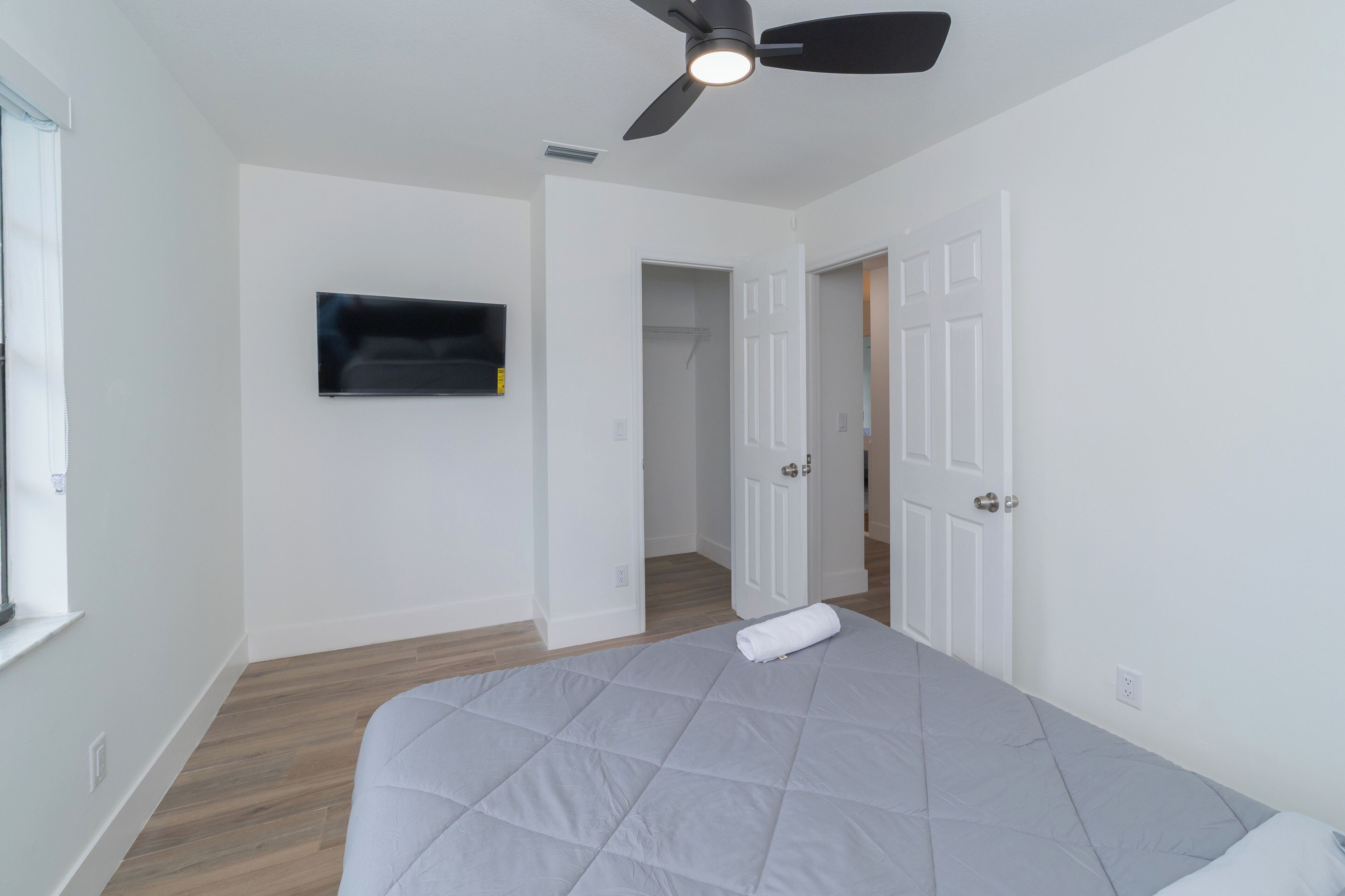 Une chambre à coucher comprenant un lit, un ventilateur de plafond, un téléviseur fixé au mur et une porte donnant sur une autre pièce.
