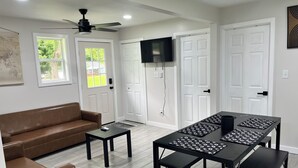 A modern living room with a brown leather sofa, a black coffee table, a dining table with black chairs, a ceiling fan, and a flat-screen TV mounted on the wall.
