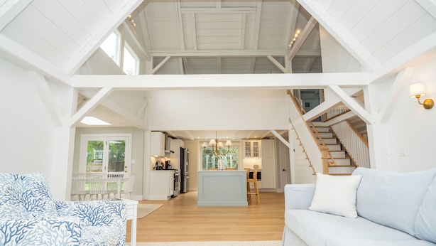 A spacious loft-style living area with a high ceiling, wooden beams, a central island with a chandelier, a staircase, and a light blue sofa. - All-New Coastal Luxury Near CBI & Beaches, Dog-Friendly (Chatham)