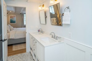 Une salle de bain moderne avec un double vasque, un grand miroir et un lit dans la pièce adjacente.