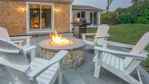 A patio with a stone fire pit surrounded by Adirondack chairs, a grill, and a house with a stone facade and windows. - All-New Coastal Luxury Near CBI & Beaches, Dog-Friendly (Chatham)