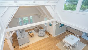 A spacious loft with a high ceiling, wooden flooring, a living area with a sofa and coffee table, a dining area with a table and chairs, and large windows offering views of greenery. - All-New Coastal Luxury Near CBI & Beaches, Dog-Friendly (Chatham)