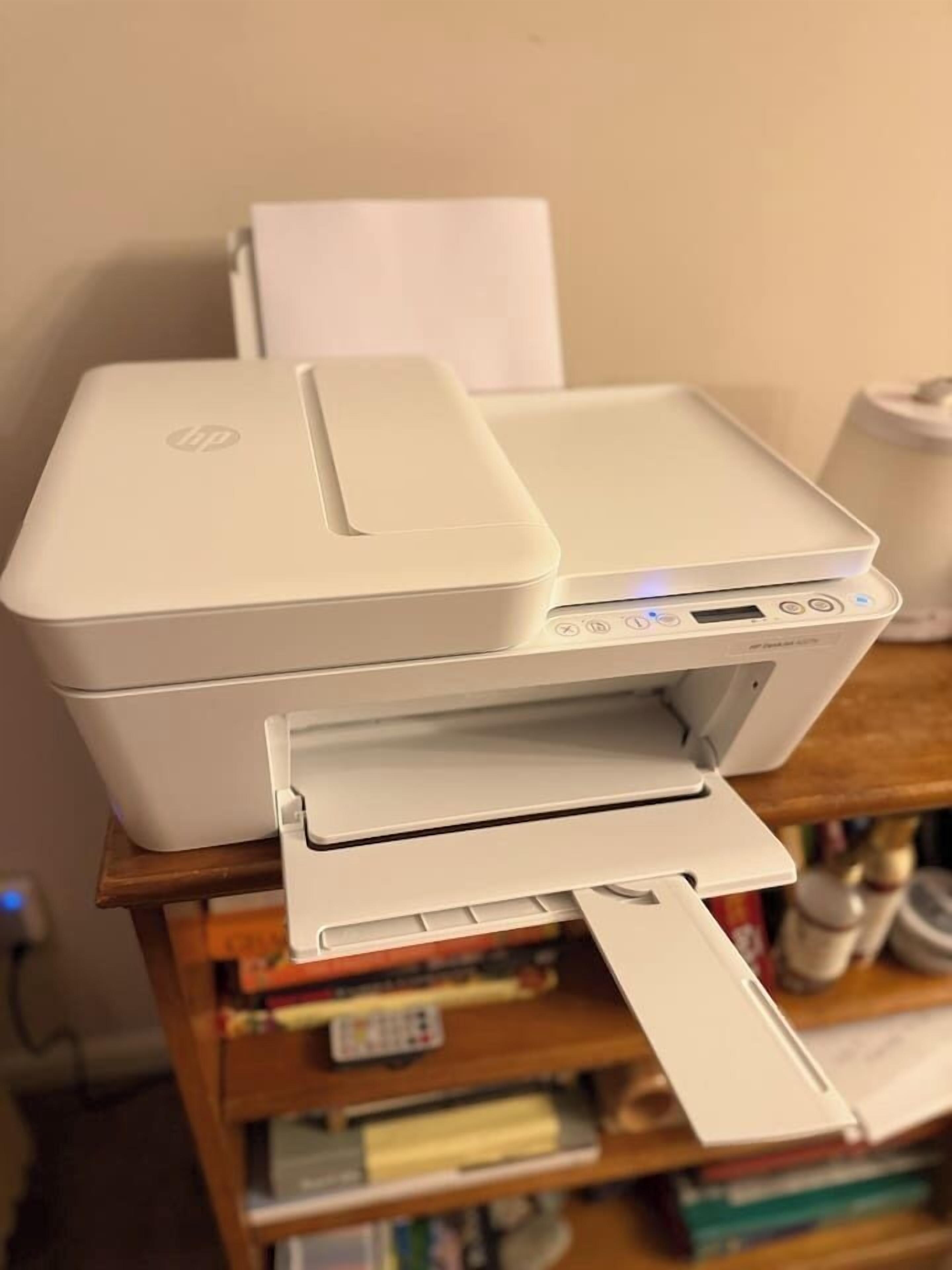 Une imprimante HP blanche est posée sur une table en bois, avec une étagère remplie de livres derrière.
