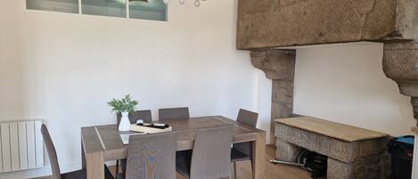 A dining area with a wooden table, six chairs, a vase with a plant, and a stone structure resembling a fireplace.