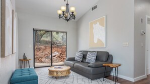 A modern living room with a grey sofa, a blue ottoman, a round coffee table, and a large window with a view of a stone wall and greenery.