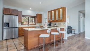 A modern kitchen with wooden cabinets, a stainless steel refrigerator, a large island with bar stools, and a window overlooking a garden.