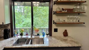 Cocina con encimera de granito, pileta de acero inoxidable y muebles de madera. Hay dos lámparas colgantes, una ventana con vista a árboles y un estante con cristalería y una botella.
