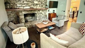 Una sala de estar acogedora con chimenea de piedra, un televisor montado en la pared y un espacio de descanso cómodo con un sofá y sillones estampados.