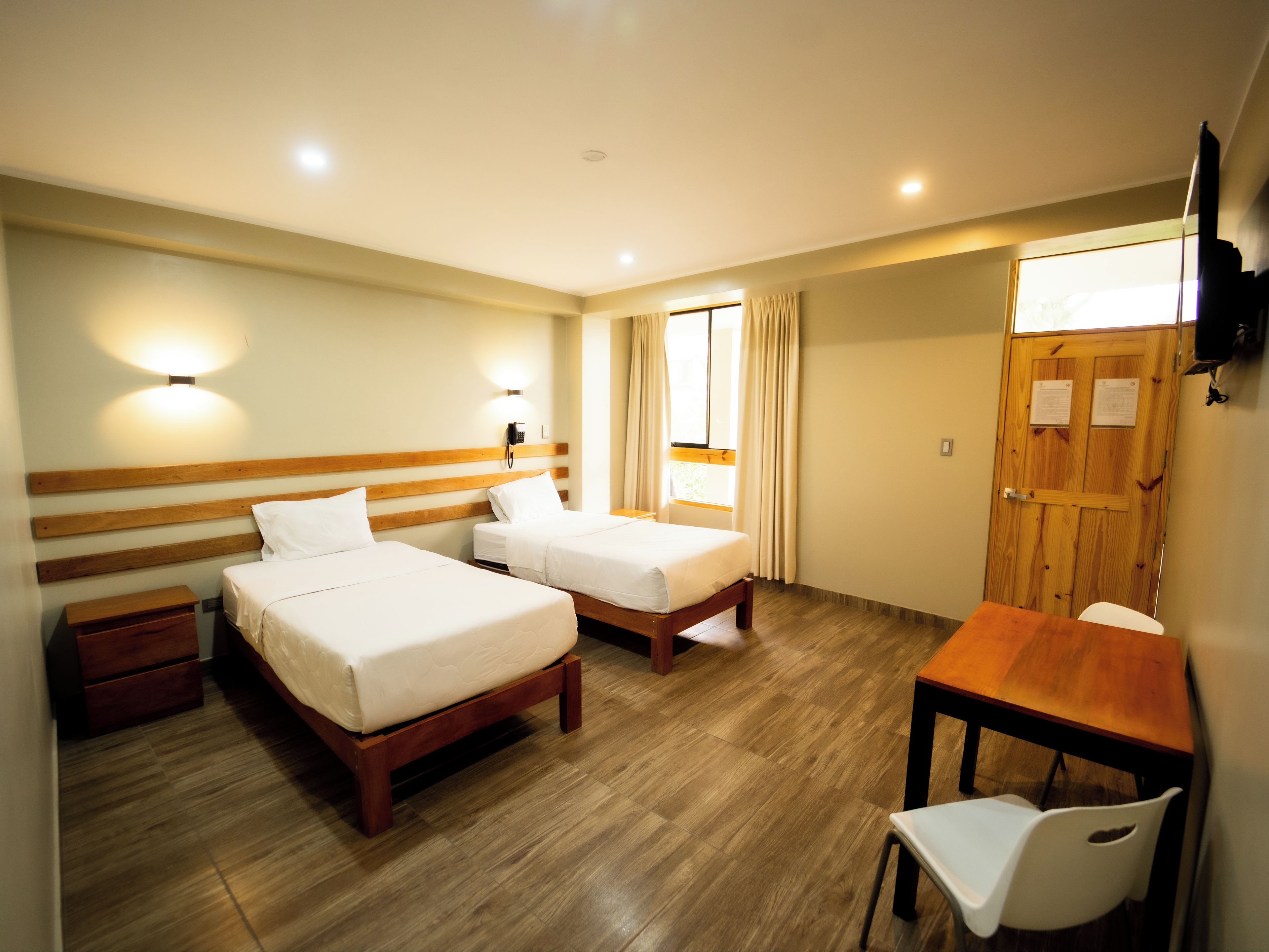 A hotel room with two single beds, a wooden desk, a chair, a nightstand, and a wooden wardrobe.