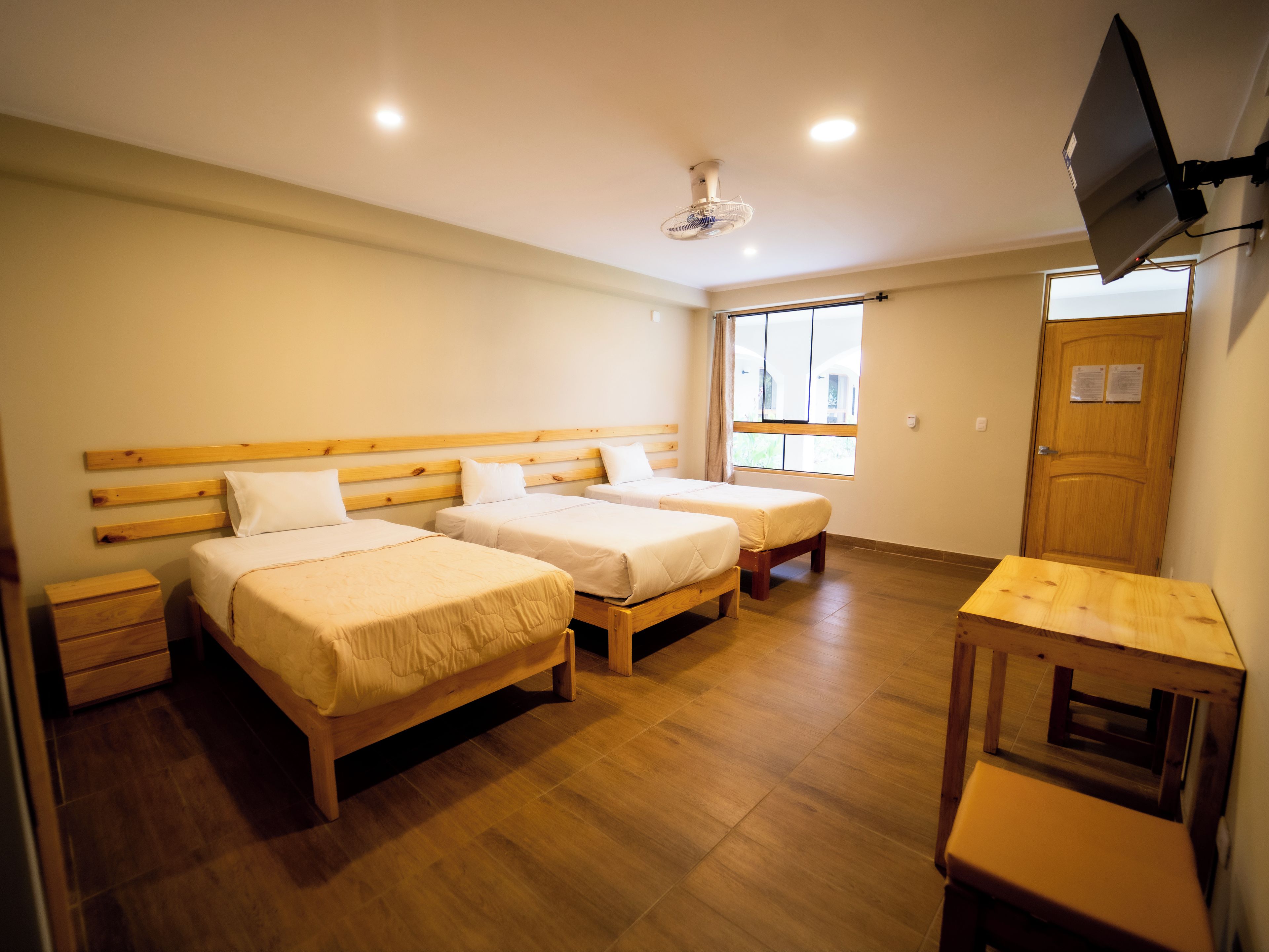 Habitación de hotel con dos camas de madera, una mesita pequeña de madera, un televisor montado en la pared y una ventana con cortinas.