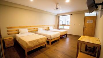 Habitación de hotel con dos camas de madera, una mesita pequeña de madera, un televisor montado en la pared y una ventana con cortinas.
