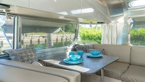 A boat interior with a dining area, cushioned seating, and a view of greenery through the windows.