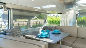 A boat interior with a dining area, cushioned seating, and a view of greenery through the windows.