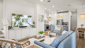 A modern living room with a large TV, white cabinetry, and a dining area with wicker furniture. - Birds Nest 160 Tai-f (Port Aransas)