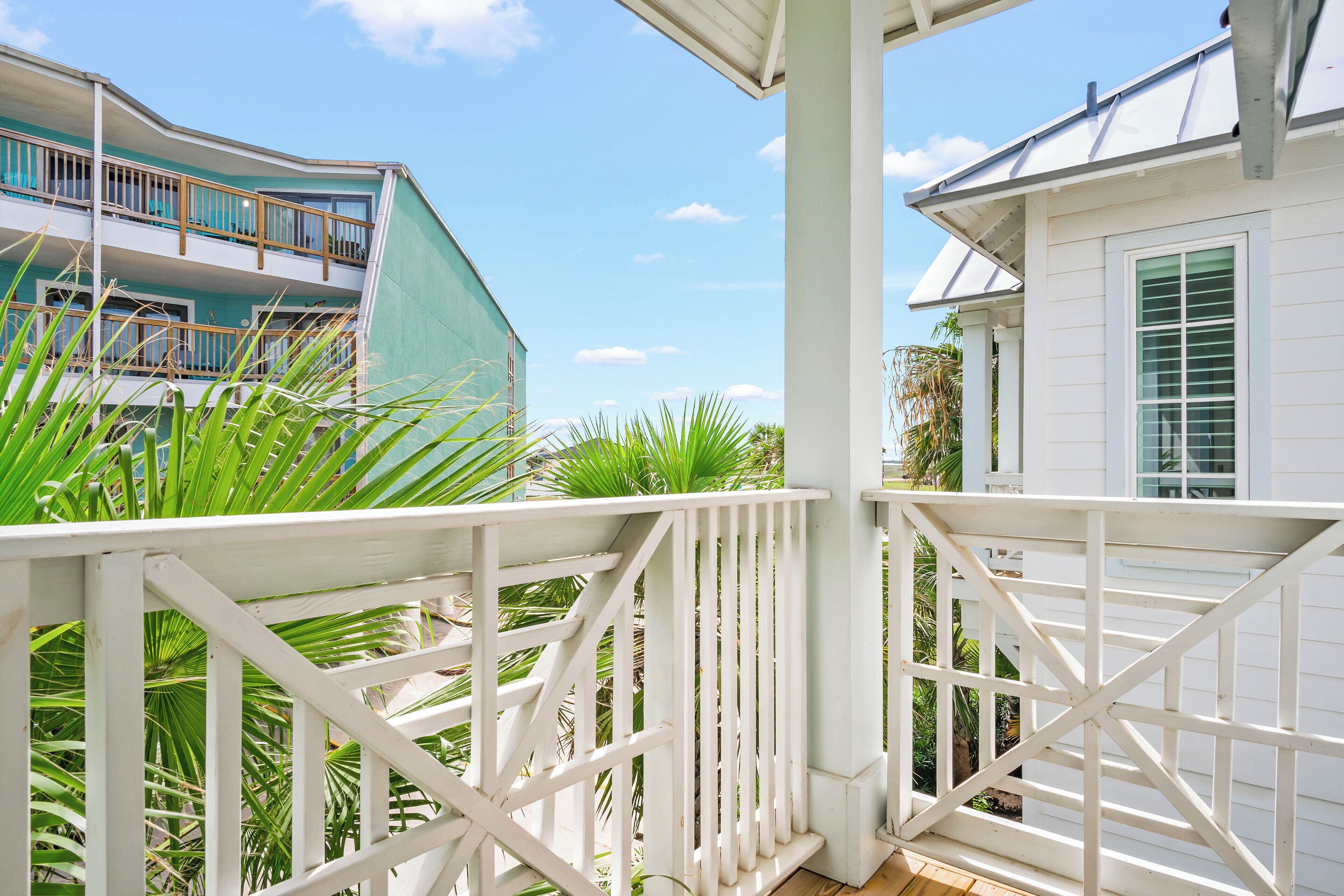 A white balcony with a white railing, overlooking a building with a green facade and a white building with a window.