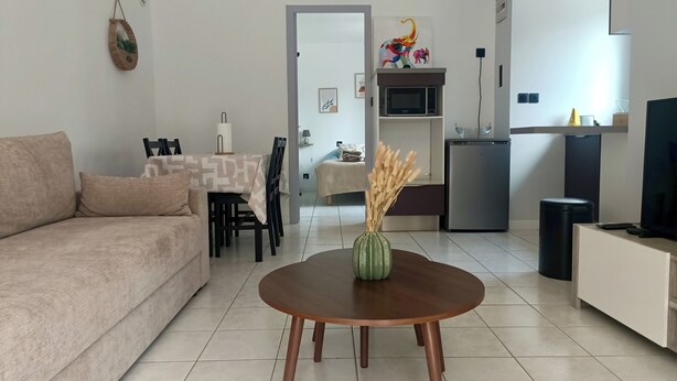 Een moderne woonkamer met een bank, een eettafel met stoelen, een keukenhoek met een koelkast en een televisie.