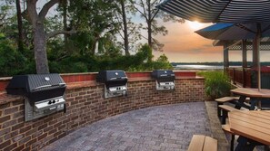 En udendørs terrasse med tre grill, et træbord og udsigt til søen i solnedgangen.