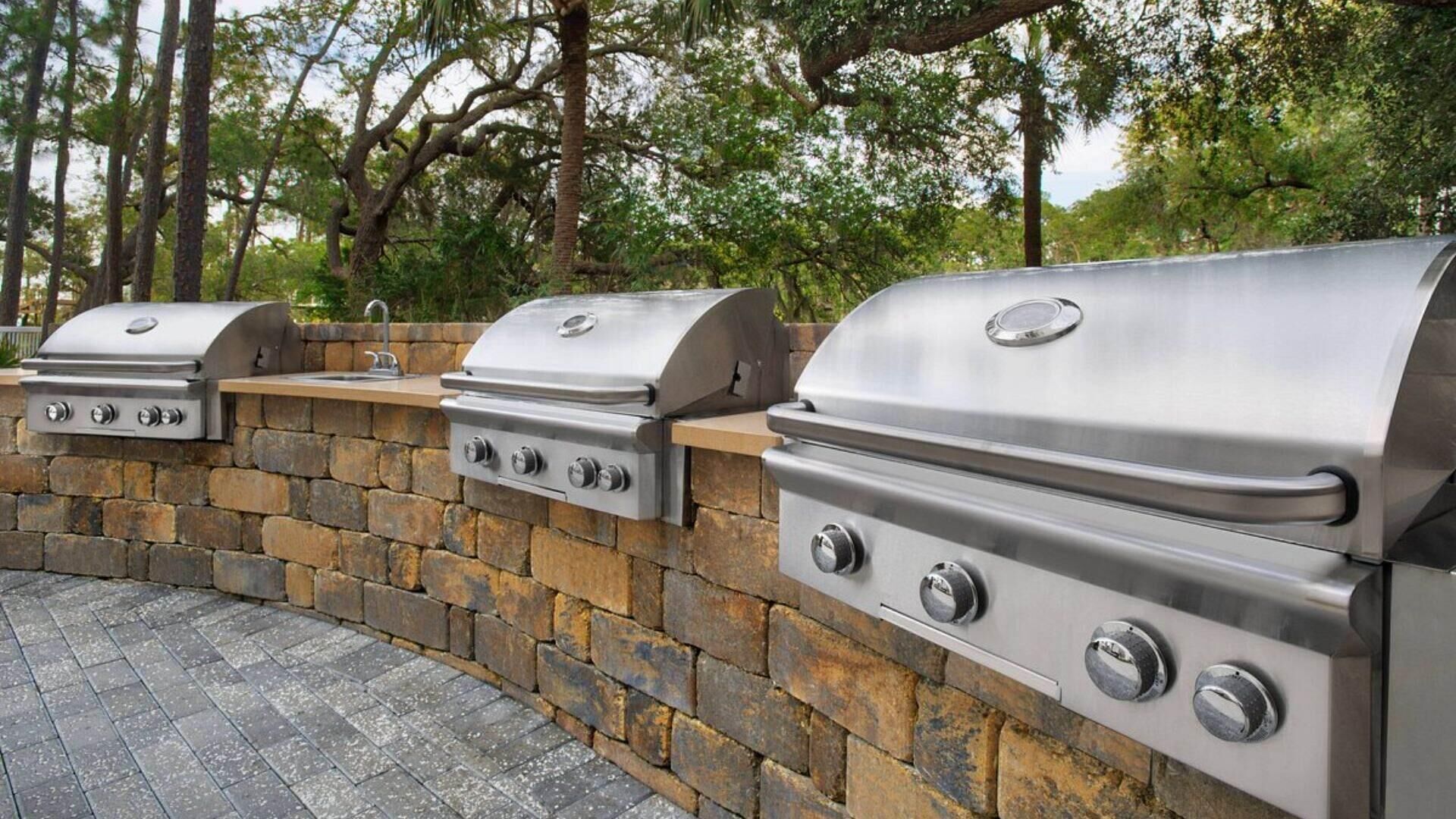Een buitenkeuken van natuursteen met meerdere roestvrijstalen barbecues, een geïntegreerde spoelbak en een pad van baksteen.
