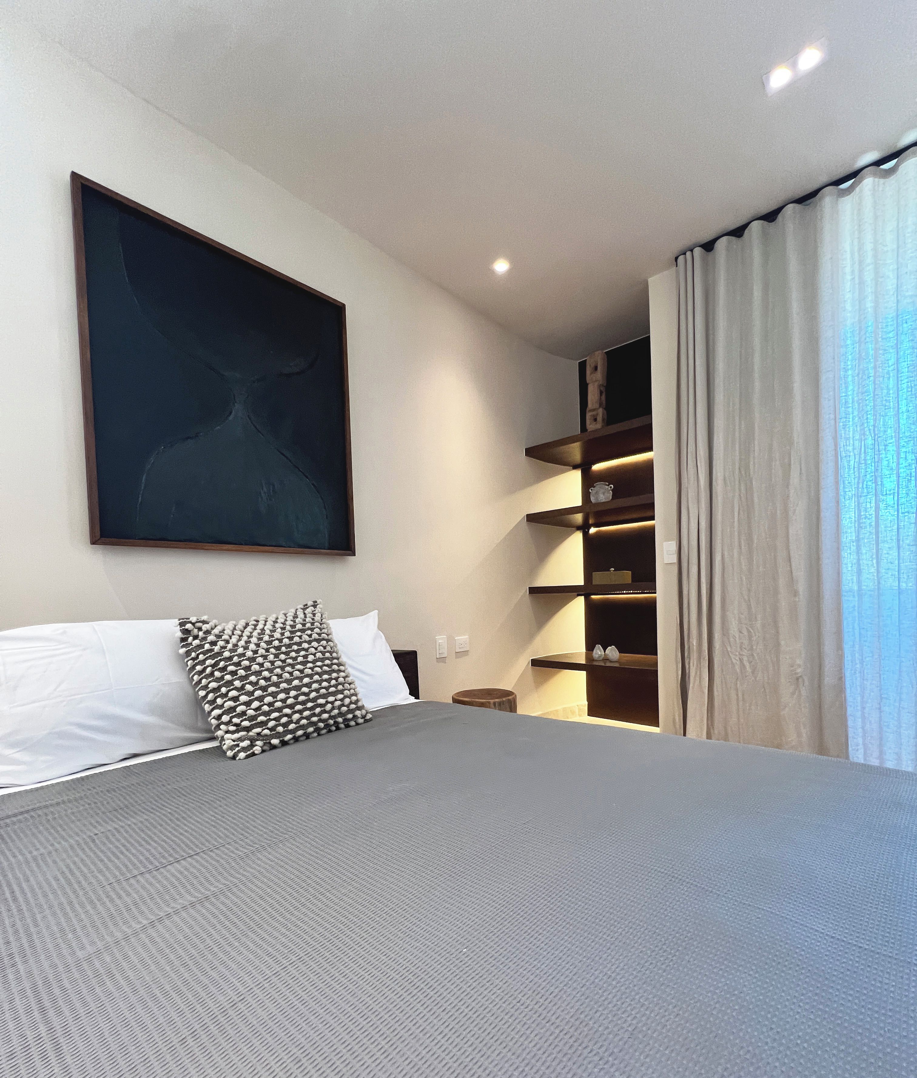 A modern hotel room with a large bed, a wall-mounted artwork, a built-in shelf with lighting, and a view of the outside through sheer curtains.