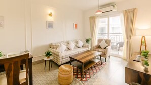 Un salon chaleureux avec un canapé, une table basse en bois et une table à manger avec des chaises. Il y a un balcon avec vue, un ventilateur de plafond et une lampe sur une table d’appoint.
