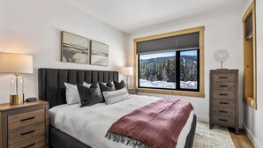 A bedroom with a large bed, two bedside tables, a window with a view of snow-covered trees, and a dresser. - Switchback Creek Condo 4 (Sun Peaks)