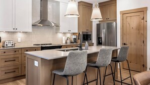 A modern kitchen with white cabinets, a stainless steel refrigerator, a central island with bar stools, and pendant lighting. - Switchback Creek Condo 4 (Sun Peaks)