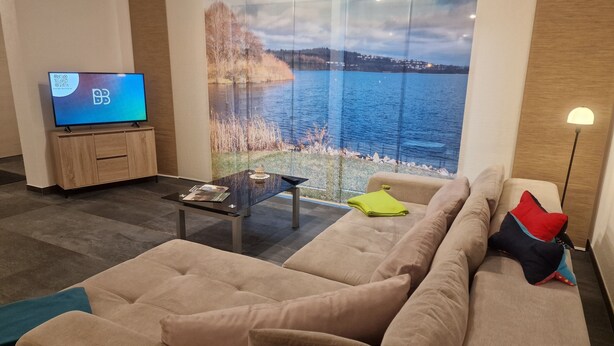 Un salon moderne doté d’une grande fenêtre donnant sur un lac, d’un téléviseur à écran plat et d’un canapé confortable agrémenté de coussins.