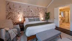 Ein Schlafzimmer mit einem großen Bett, Nachttischen, einem Sessel und einer Bank. Eine Tür führt zu einem Badezimmer mit Dusche und Waschbecken.