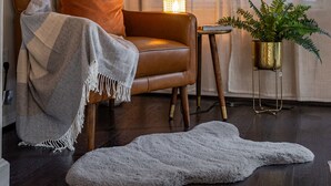 Ein gemütliches Wohnzimmer mit Ledersessel, einer großen Topfpflanze, einem kleinen Tisch mit Lampe und einem grauen Teppich auf Holzdielen.