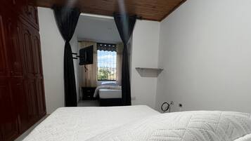 Un dormitorio con cama, techo de madera, paredes blancas, ventana con cortinas y televisor montado en la pared.
