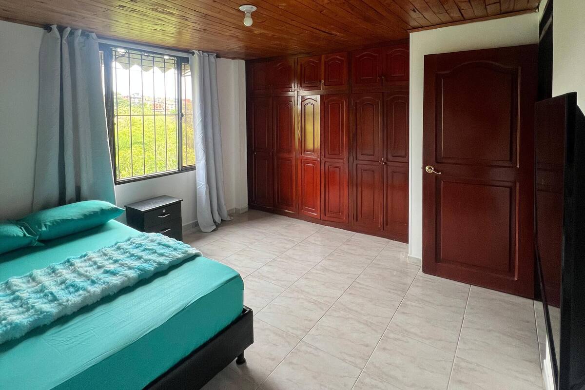 Un dormitorio con techo de madera, cama con colcha color turquesa, ventana con cortinas y un armario.