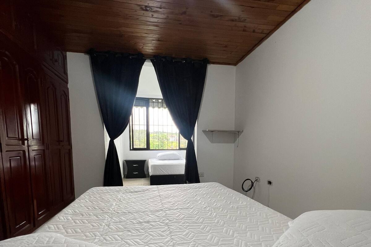 Un dormitorio con una cama grande, techo de madera, una ventana con cortinas y una mesita de noche.