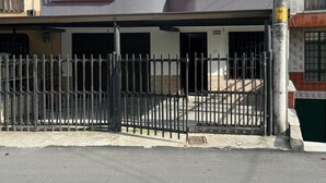Un edificio de dos pisos con entrada protegida por un portón, puerta de garaje visible y un poste de electricidad con cables.