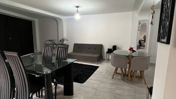 A modern living room with a dining area, featuring a glass-top table, grey sofas, and a black rug.