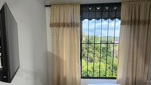 Un dormitorio con cama, mesita de noche, televisor montado en la pared y ventana con cortinas.
