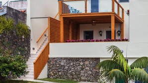 A modern two-story building with a wooden staircase, a balcony with outdoor seating, and a stone wall.
