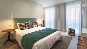 A bedroom with a bed, a green quilt, two bedside tables, a chair, a window with curtains, and a small stool.