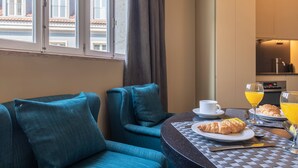 Et moderne hotelværelse med spiseplads, der inkluderer et rundt bord, en blå sofa samt morgenmadsvarer såsom croissanter, appelsinjuice og en kop.