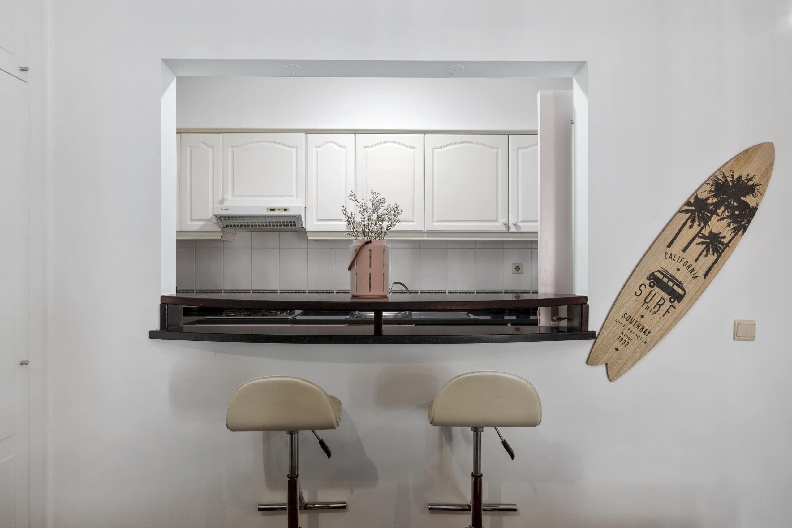 Een keuken met witte kasten, een roze vaas met bloemen en een surfboard decoratie.
