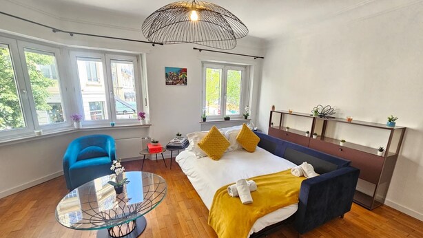 A modern bedroom with a bed, a blue chair, a glass table, and a large window. - Ideal city stay 1BR near train station (Luxembourg City)