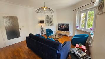 Ein Wohnzimmer mit einem blauen Sofa, einem Glastisch, einem blauen Sessel, einem Fernseher und einem Gemälde an der Wand.